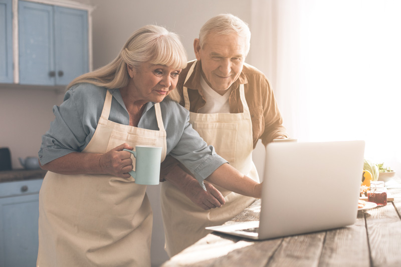 Excited senior man and woman are reading interesting recipe on laptop and smiling. They are standing in kitchen and drinking tea. Couple is wearing chef aprons